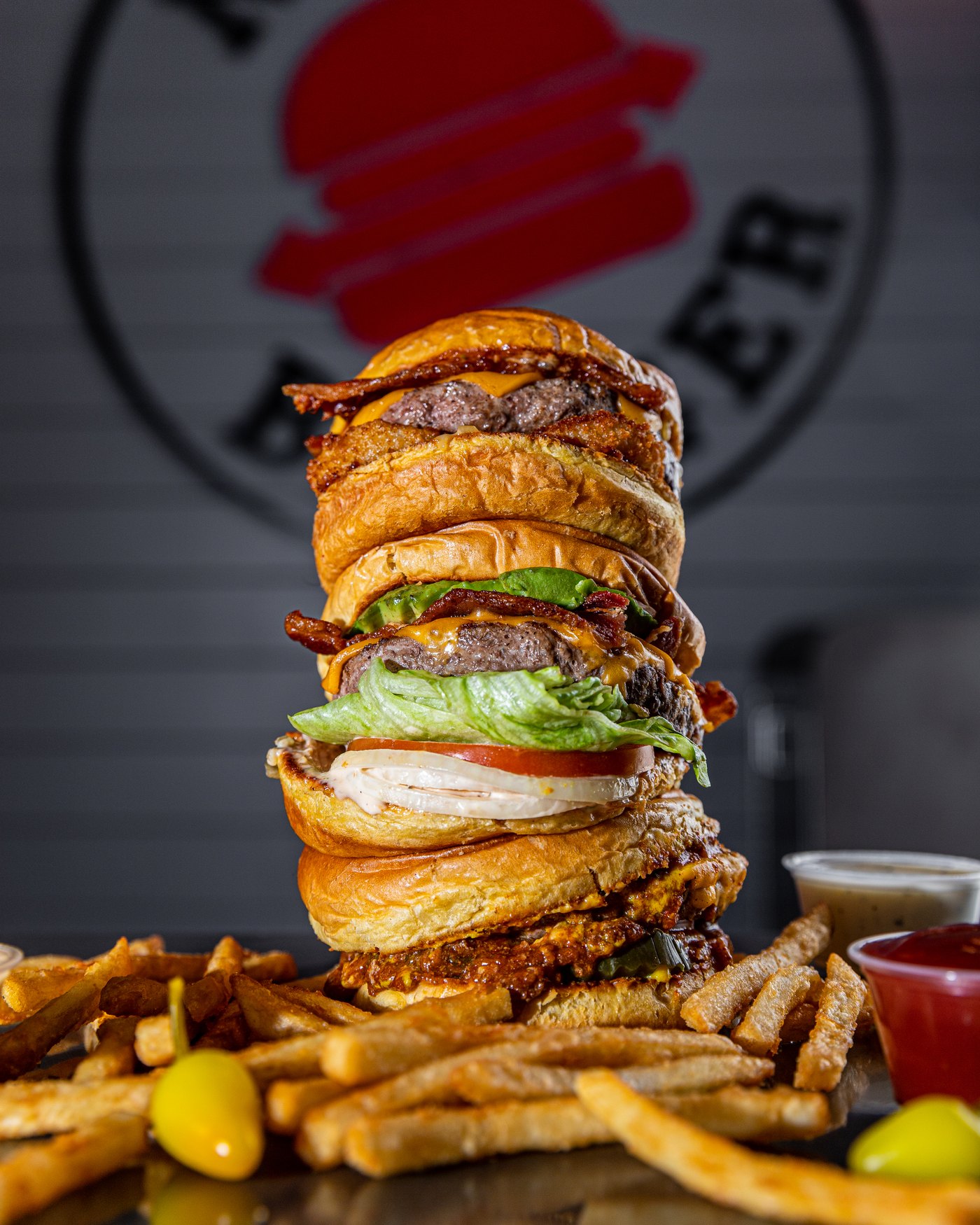 A stack of Nexx Burgers with fries, chili peppers and dipping sauces, in front of the Nexx Burger logo